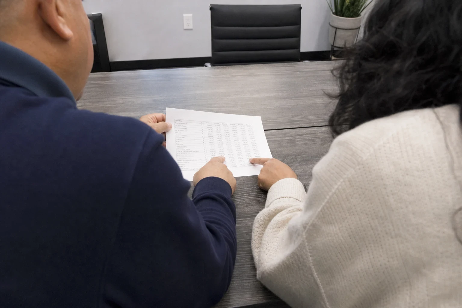 Over-the-shoulder view of two people at a gray conference table reviewing a printed page of figures, one pointing at a column, with an empty office chair beyond.