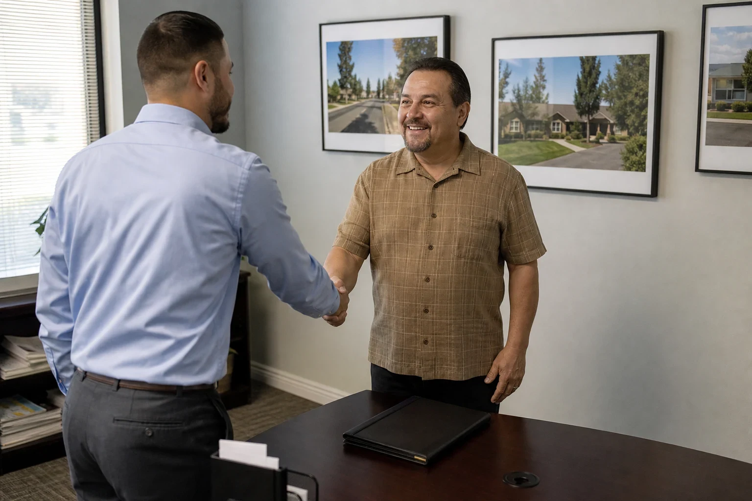 Insurance agent in a blue shirt shakes hands with a smiling client beside a dark desk with a closed folder, framed neighborhood photos on the office wall