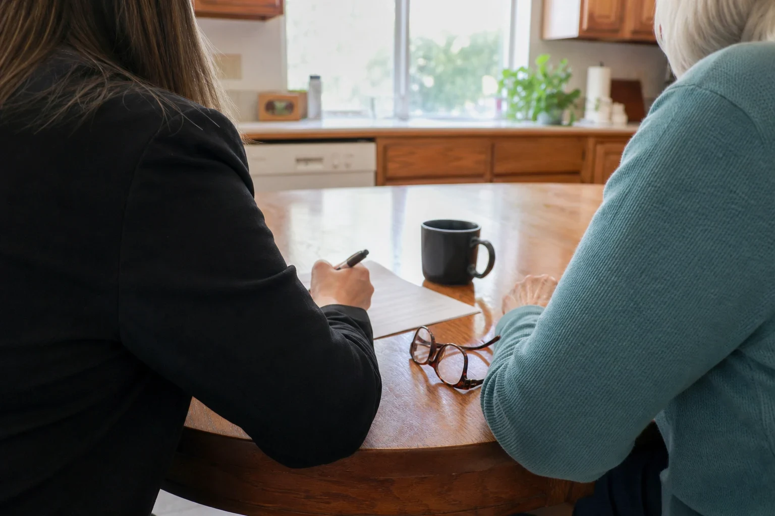 Agent in a dark blazer writes on a paper at a round wooden kitchen table beside an older woman in a teal sweater, with glasses and a black mug on the table