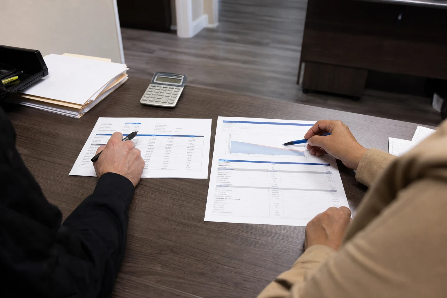 Two people at an office desk, hands holding pens over a financial statement and a coverage chart laid side by side, with a calculator and file folders nearby